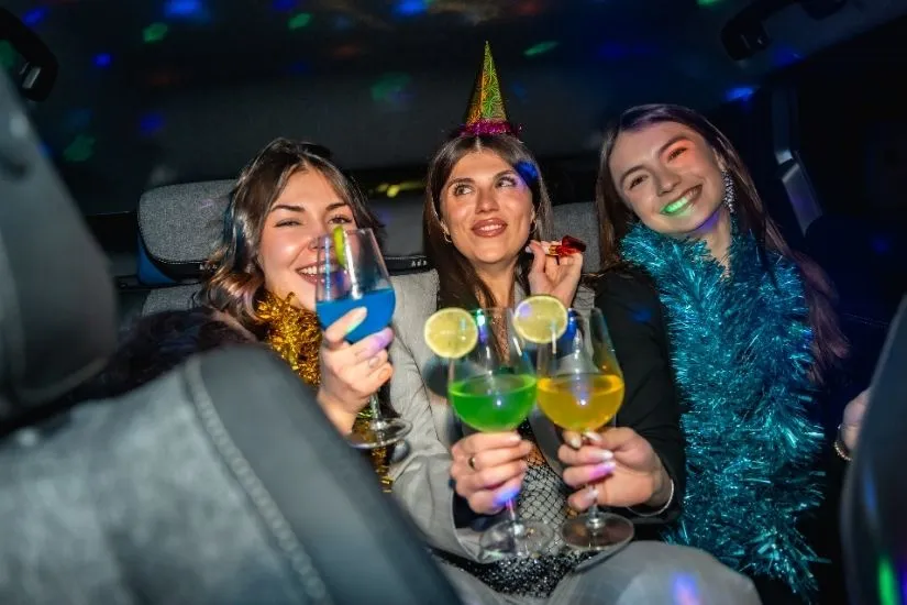 hree friends in party hats and tinsel celebrating a birthday with colorful drinks in the back of a car with disco lights.